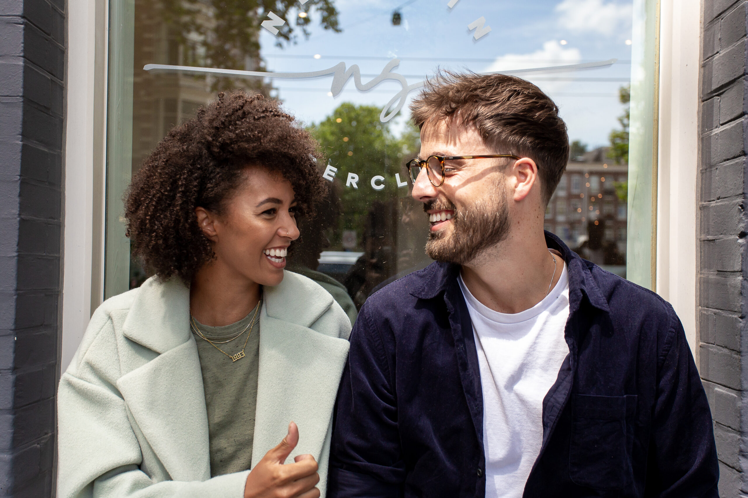 Blij patiënten-koppel dat elkaar lachend aankijkt voor de ingang van New Skin Laserclinic Amsterdam.